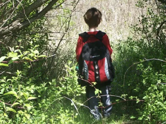 A first-grade student with a backpack walks through the woods.