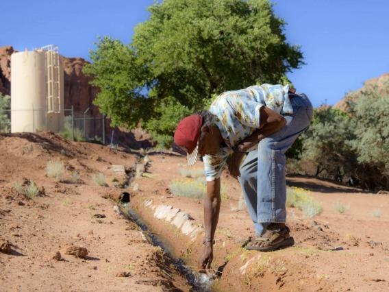 A man bends to check an irrigation pipeline in dry soil on a sunny day.