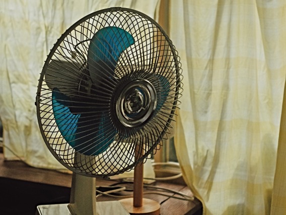 A blue electric fan in front of a beige curtain.