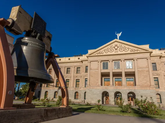 The Arizona liberty bell is in the foreground with the white Arizona state capitol building in the background, with the winged Victory statue atop.