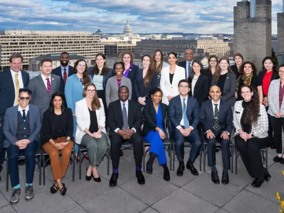 A group of men and women in professional attire smiling outdoors. White brick buildings are in the background.