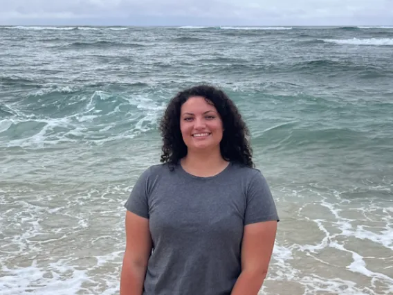 Caitlin Brady: Woman with dark curly hair at the beach with ocean in the background. 