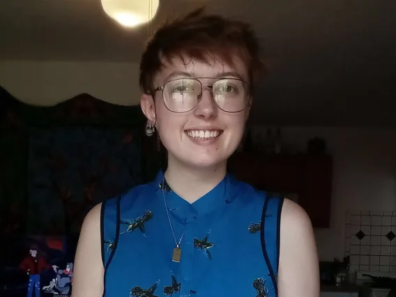 Emery Byrde: Person with short brown hair and wire-frame classes wearing a blue  hummingbird-print top smiling at camera