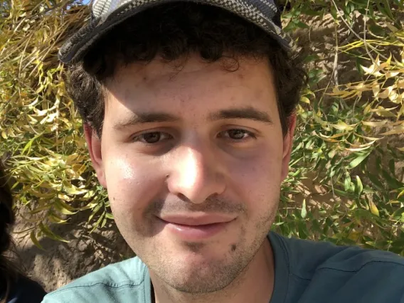 Myles Kashanipour: Man with dark curly hear wearing a grey hat and teal shirt. Background is a bush or a tree close up.