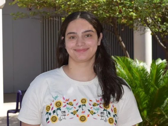 Nadera Alami: Woman with long dark hair wearing a patterned white t-shirt. She is standing in front of some trees