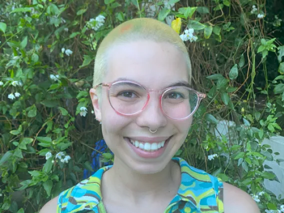 Riley Remen: Person with buzzed yellow hair wearing a green-patterned tank top and round pink glasses smiling at camera. They are standing in front of some vines.