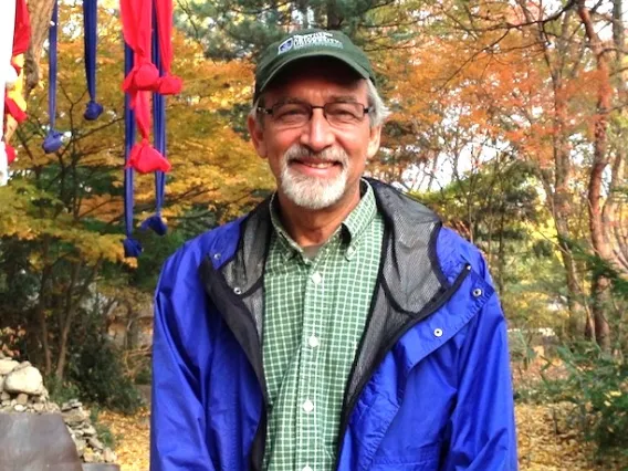 Larry Fisher, gentleman wearing plaid green shirt, blue jacket, and a green baseball hat surrounded by trees in fall colors.