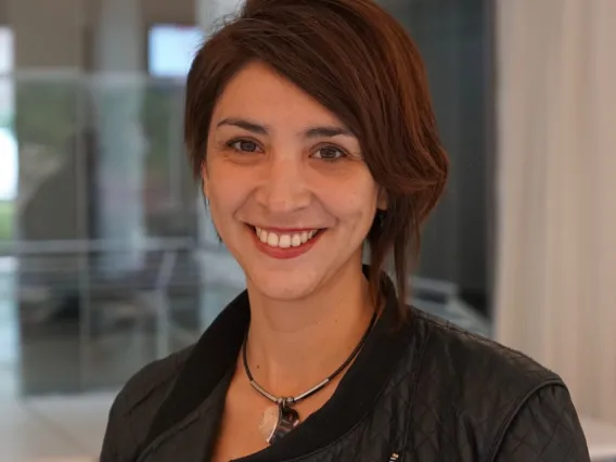 Carolina Heredia, young woman smiling with short brown hair swept to one side wearing black jacket. Standing in front of reflective glass background.