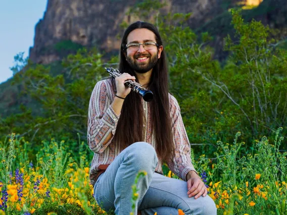 Chris Zatarain, man with long brown hair crouched in wildflowers holding an oboe in front of mountain.