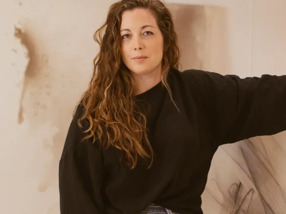 Heather Bird Harris, woman with long brown curly hair wearing a black sweater and blue jeans in front of a beige colored artistic background.