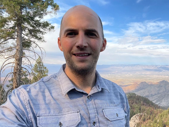 Philip Alejo on a mountain with pine tree, landscape, and cloudy blue sky in the background