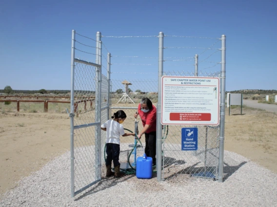 A woman and child collect water from a faucet into buckets. The faucet is protected by chain link fence in a desert.