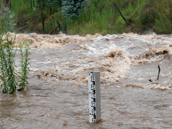 A flooding river rages behind a stick showing the water height.