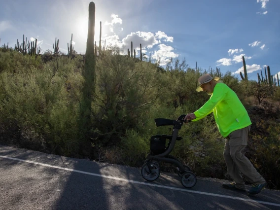 A man in a hat and green jacket pushes a walker uphill in the afternoon sun.