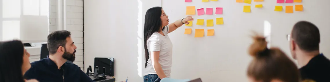 a group of people in an office setting watching as someone stands in front of them, gesturing to multiple sticky notes on the wall.