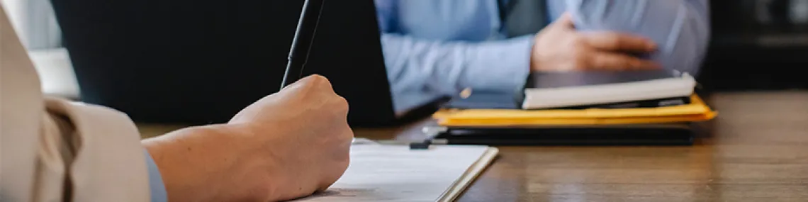 Person writing on a small stack of papers with a pen while someone sitting across the table from them.