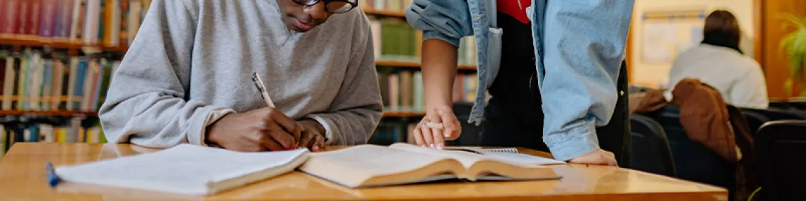two people looking at a book on a table inside a library. one of them is looking down and writing.