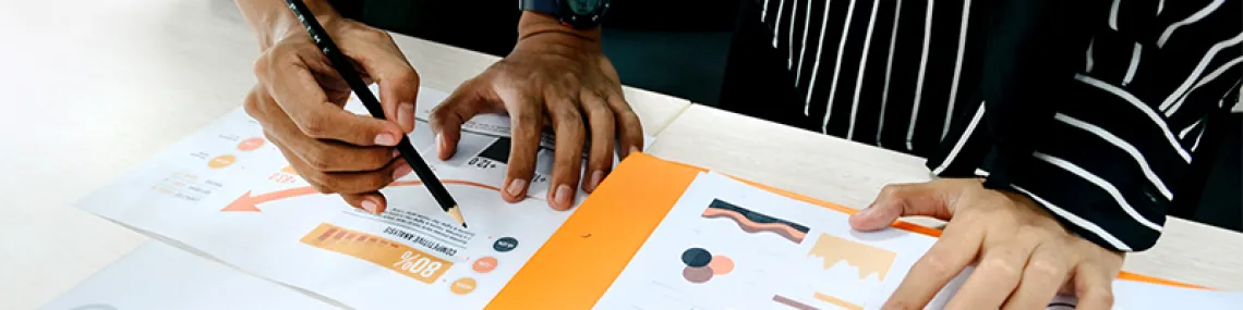 Two individuals looking down at project documents together with their hands on the flat surface. One of them is tracing an element of the document. 