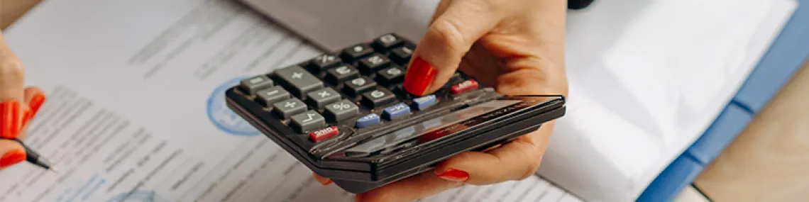 A hand holding a calculator while reviewing documents with a pen in the other hand.