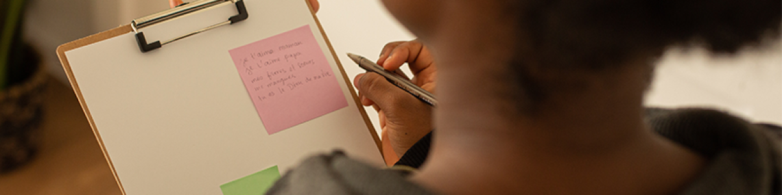 A person holding a clipboard with sticky notes on it. There is a pen in their hand and the view is from behind the person. 