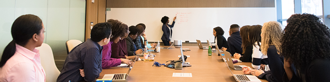 People gathered around a long table in a conference room, looking toward a person explaining something on a whiteboard.