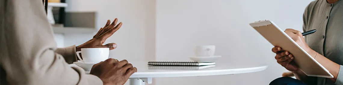 A pair of people are sitting. To the left, one person is talking with hand gestures. The person to the right is taking notes.