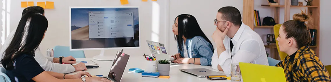 A group of people gathered around a table in an office setting. They are all looking at the screen of a computer monitor.