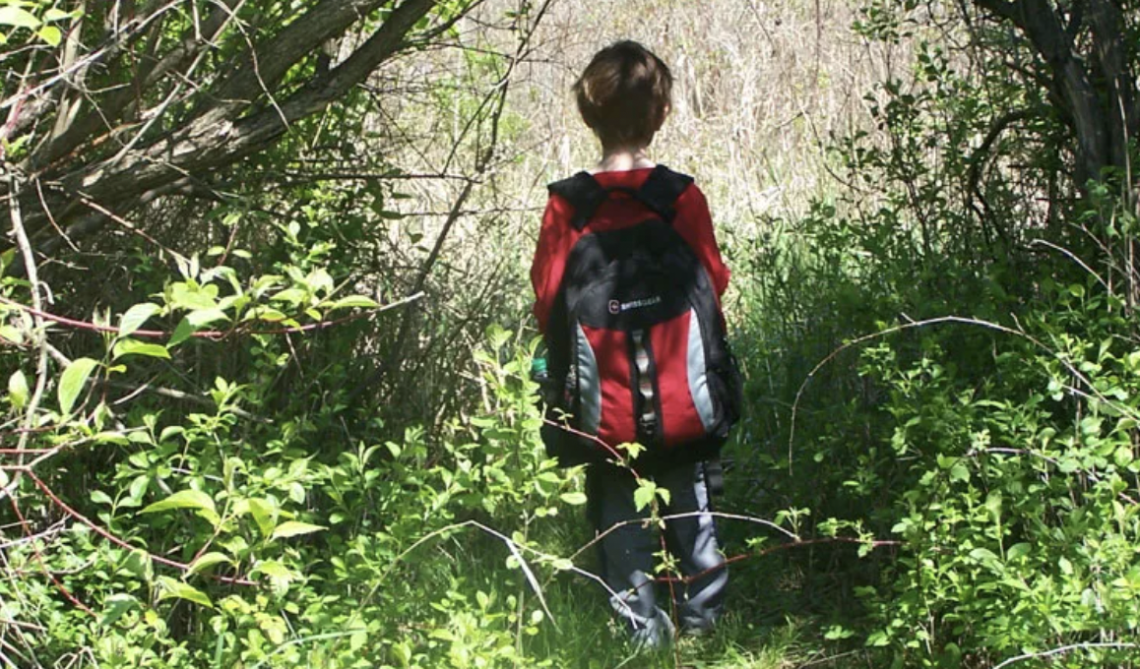 A first-grade student with a backpack walks through the woods.