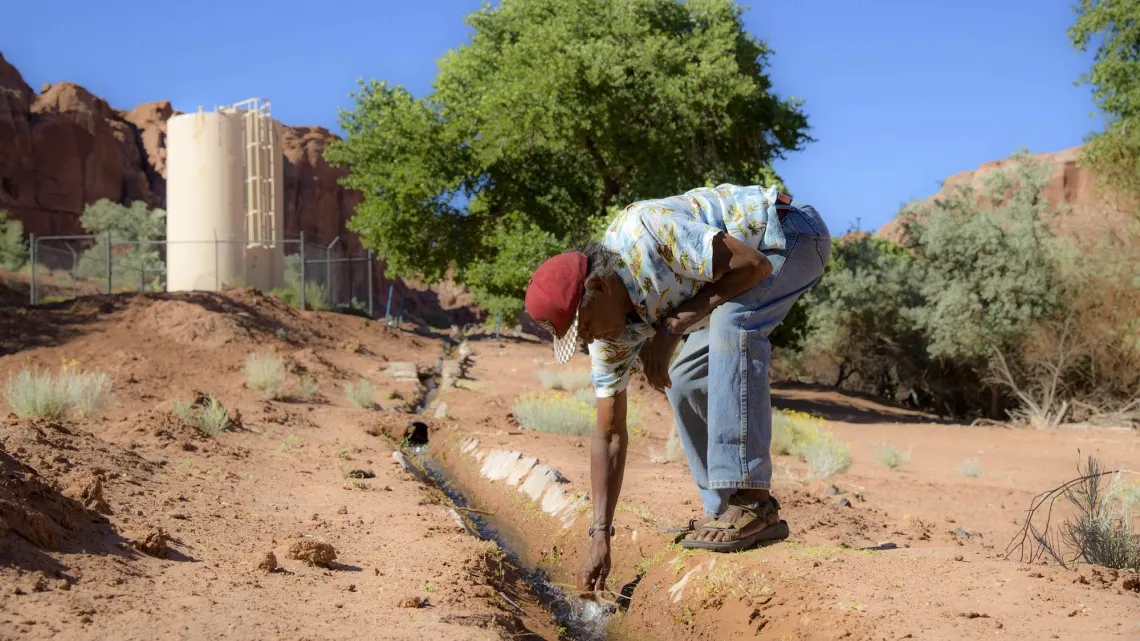 A man bends to check an irrigation pipeline in dry soil on a sunny day.