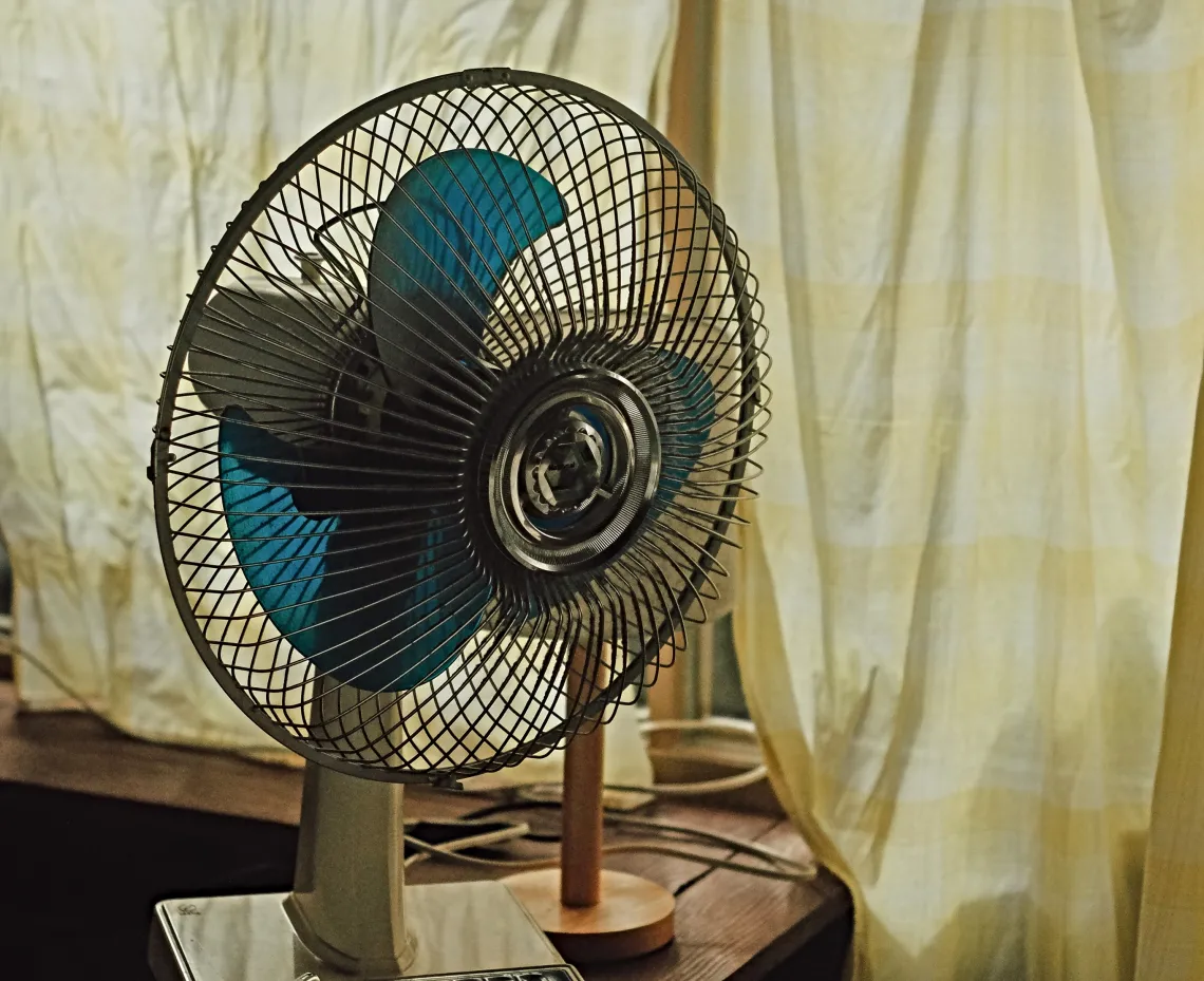 A blue electric fan in front of a beige curtain.