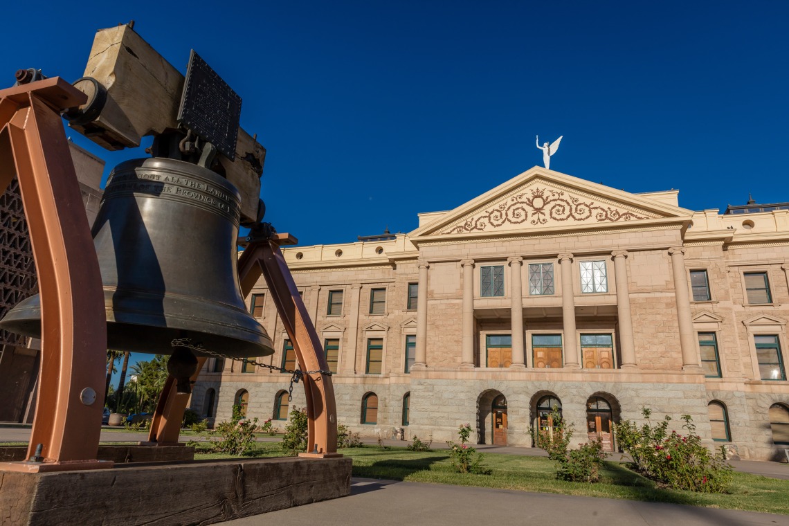 The Arizona liberty bell is in the foreground with the white Arizona state capitol building in the background, with the winged Victory statue atop.