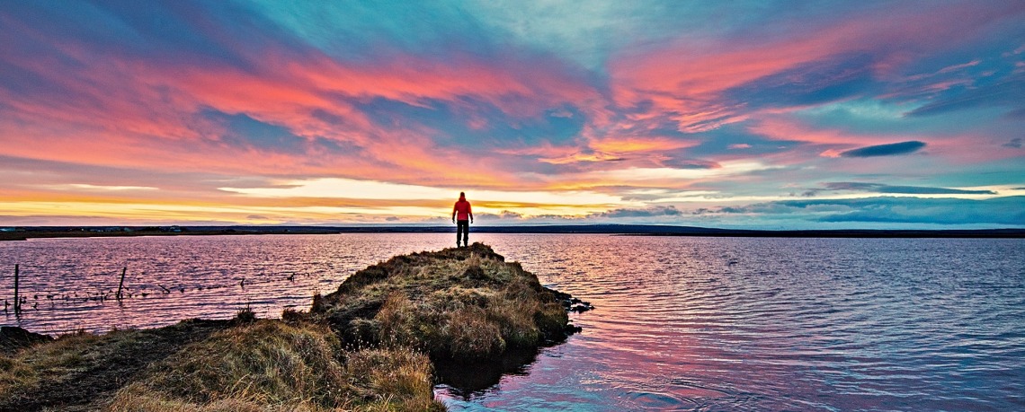 A person in a red coat stands at the end of a spit of land looking at light change over water.