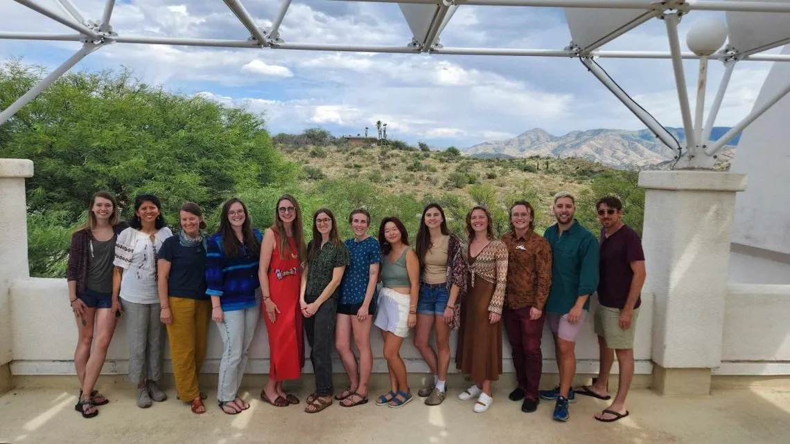 A group of young men and women are smiling together facing the camera, outdoors with mountains and clouds behind them.