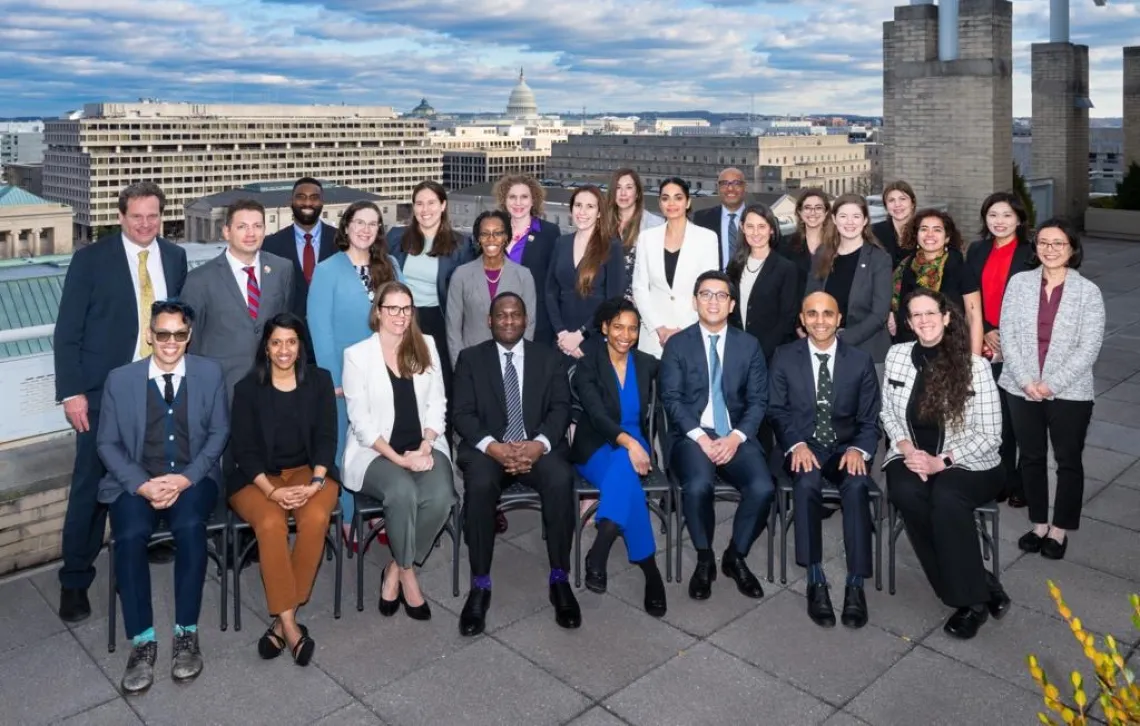 A group of men and women in professional attire smiling outdoors. White brick buildings are in the background.