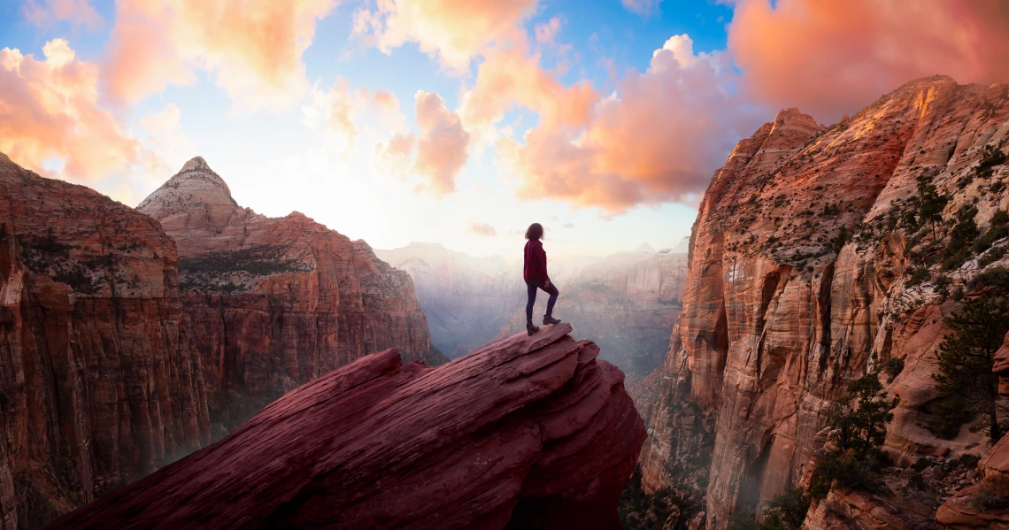 A person stands on a rocky outcrop amid mountains under a colorful sky.