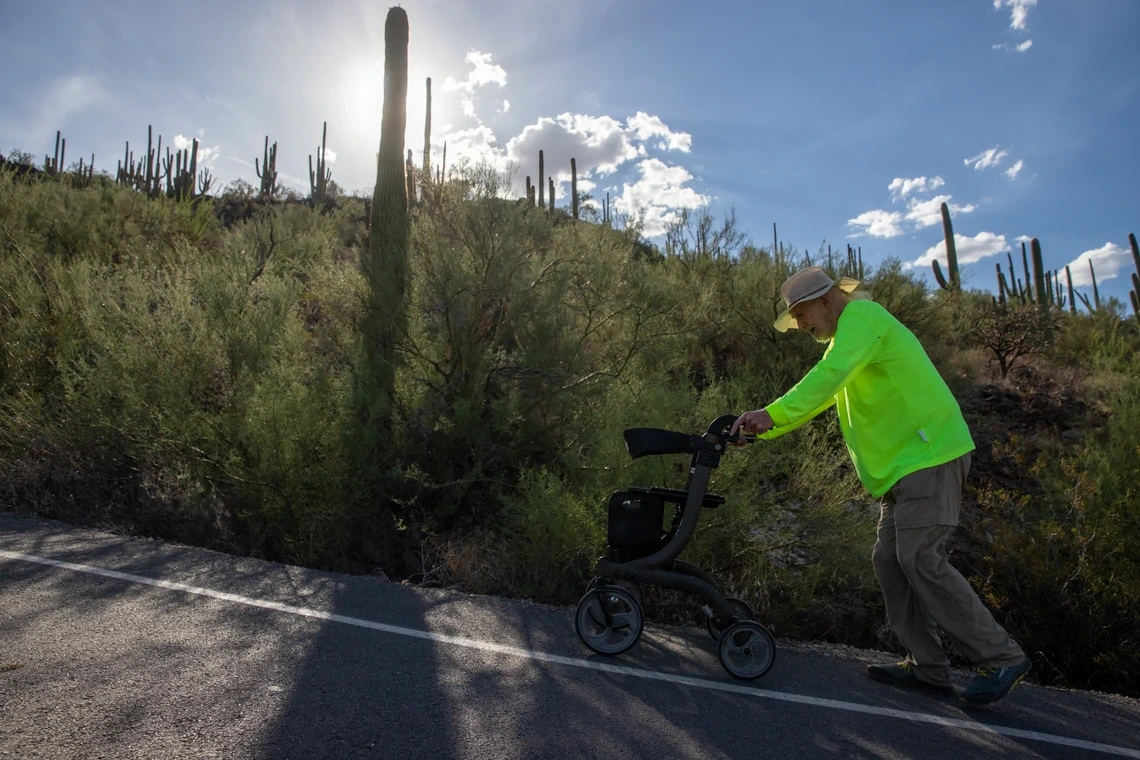 A man in a hat and green jacket pushes a walker uphill in the afternoon sun.