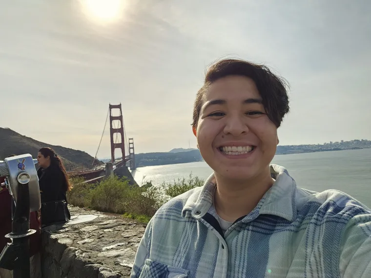 Brandolyn Baeza: Person with short brown hair wearing a blue plaid button-up smiling in front of a bridge and body of water