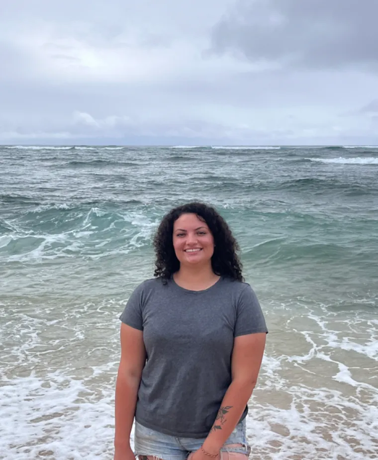 Caitlin Brady: Woman with dark curly hair at the beach with ocean in the background. 