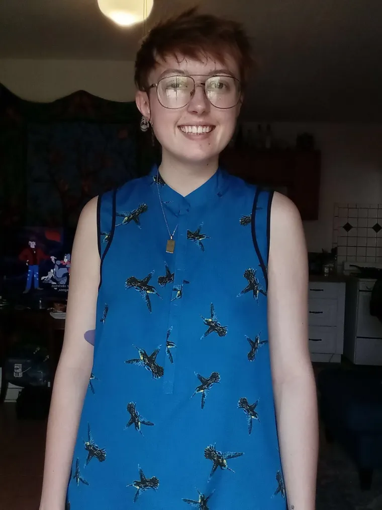 Emery Byrde: Person with short brown hair and wire-frame classes wearing a blue  hummingbird-print top smiling at camera