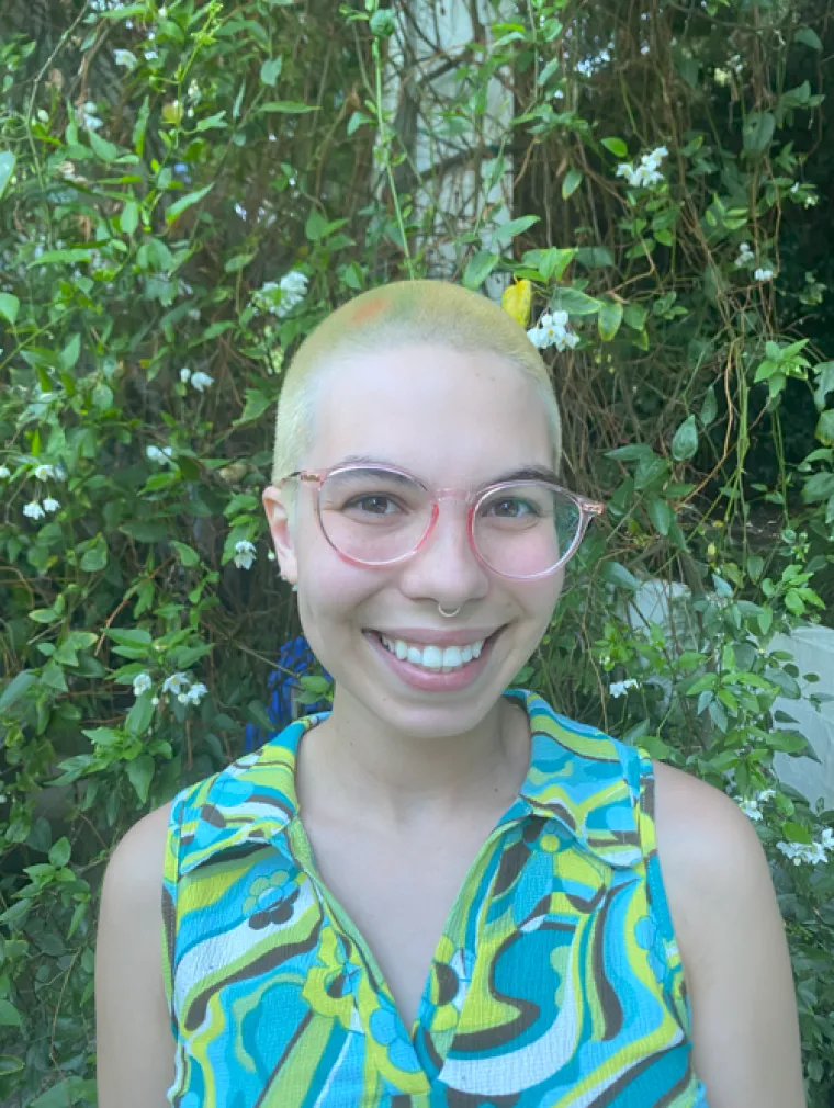 Riley Remen: Person with buzzed yellow hair wearing a green-patterned tank top and round pink glasses smiling at camera. They are standing in front of some vines.