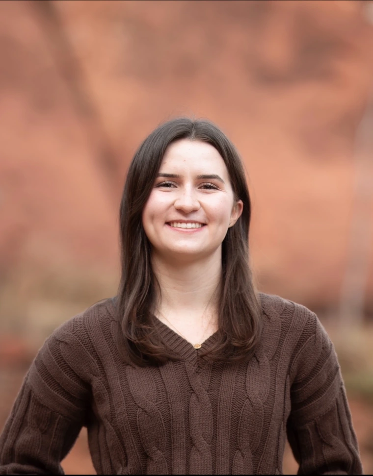 Woman with collarbone-length brown hair smiles at the camera in front of a blurry, red-rock background. She is wearing a brown knitted sweater and a small gold pendant around her neck