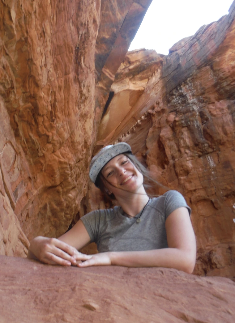 A woman with long brown hair wearing a grey baseball cap and grey shirt smiles at the camera. She is standing among a large red rock formation. Only the upper part of her body is visible. 