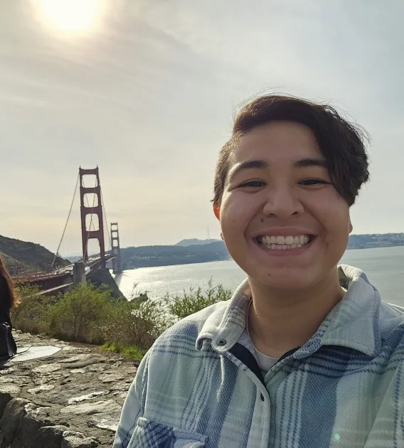 Brandolyn Baeza: Person with short brown hair wearing a blue plaid button-up smiling in front of a bridge and body of water