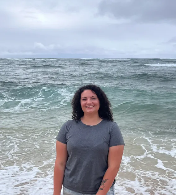 Caitlin Brady: Woman with dark curly hair at the beach with ocean in the background. 