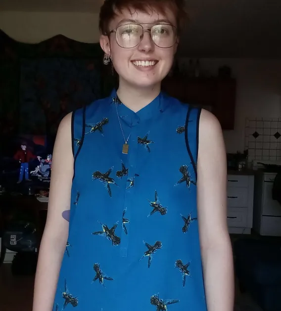 Emery Byrde: Person with short brown hair and wire-frame classes wearing a blue  hummingbird-print top smiling at camera