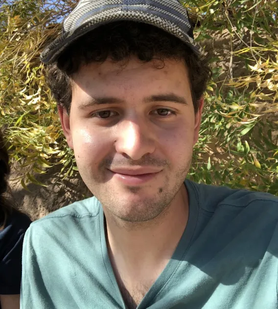 Myles Kashanipour: Man with dark curly hear wearing a grey hat and teal shirt. Background is a bush or a tree close up.