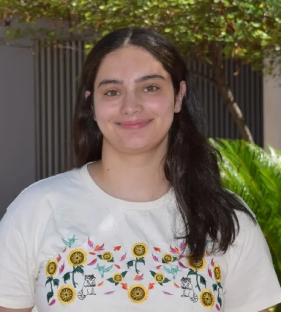 Nadera Alami: Woman with long dark hair wearing a patterned white t-shirt. She is standing in front of some trees