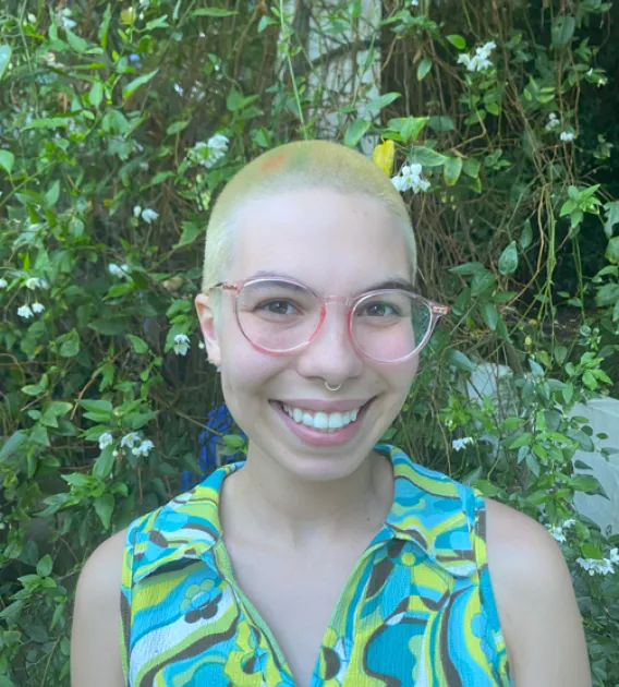 Riley Remen: Person with buzzed yellow hair wearing a green-patterned tank top and round pink glasses smiling at camera. They are standing in front of some vines.