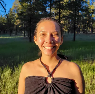 A woman smiling in a field facing the sun, wearing a black sleeveless dress and a nosering.