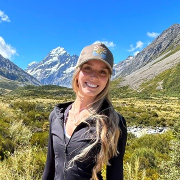 A woman in a baseball cap standing in front of a grassy valley between snow-capped mountains
